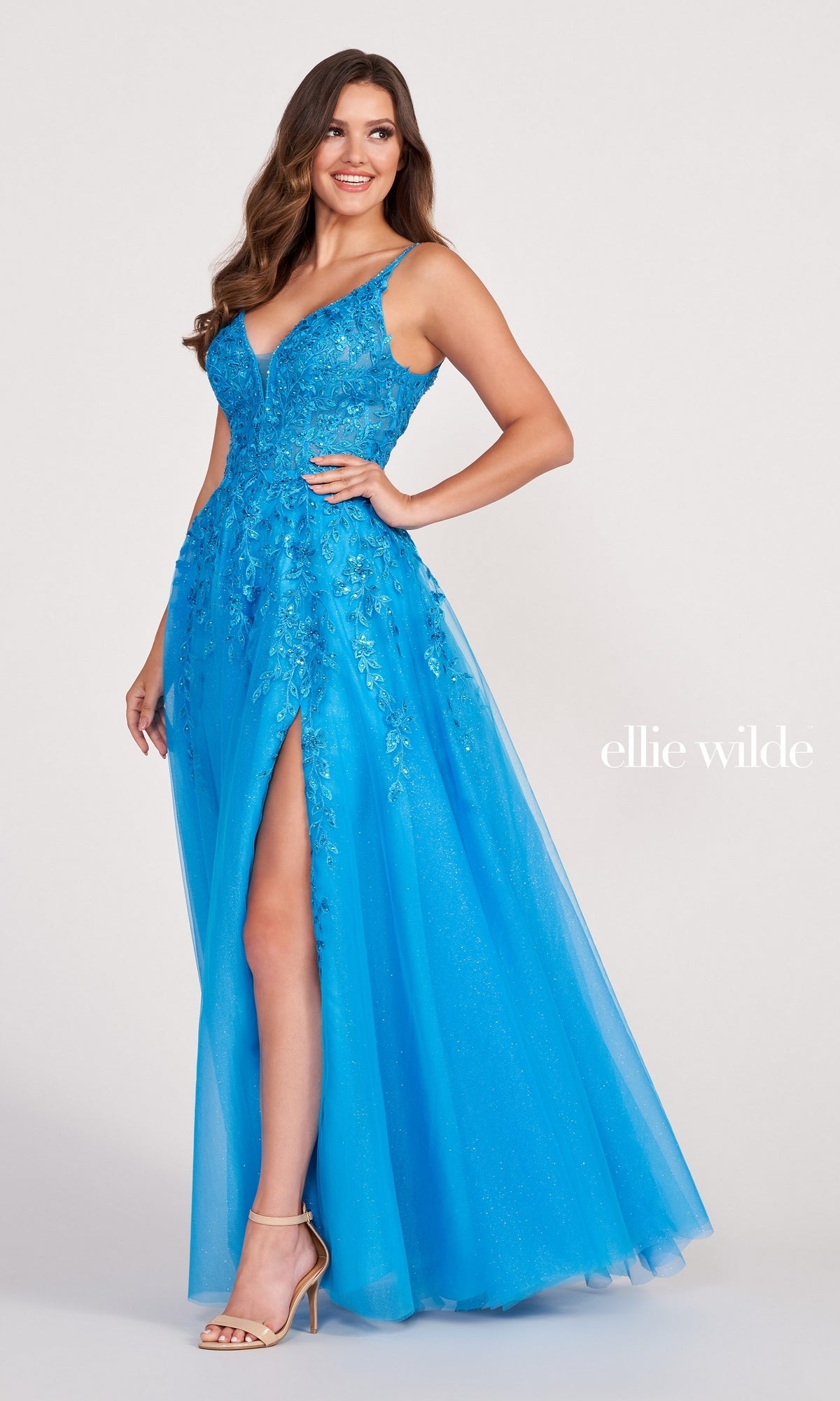 A woman poses in the Glitter-Tulle Long Ellie Wilde Prom Dress EW34089, a bright blue gown with crystal beading, A-line silhouette, high slit, and V-neckline. She pairs it with nude heels against a plain background.